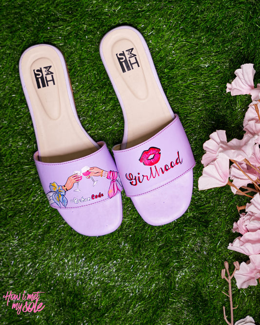 Purple slippers with 'Girlhood' text and pink flowers on a green surface