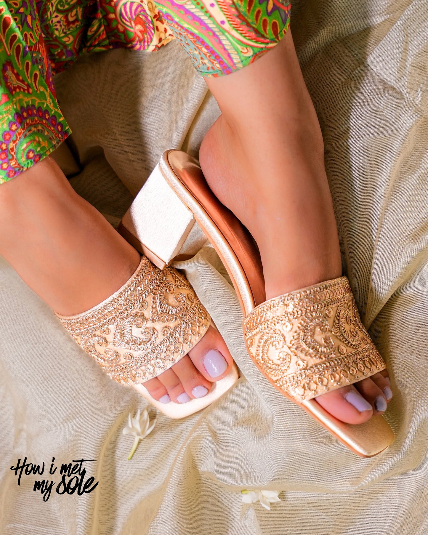 Gold embellished open toe heels worn with colorful pants on a neutral background