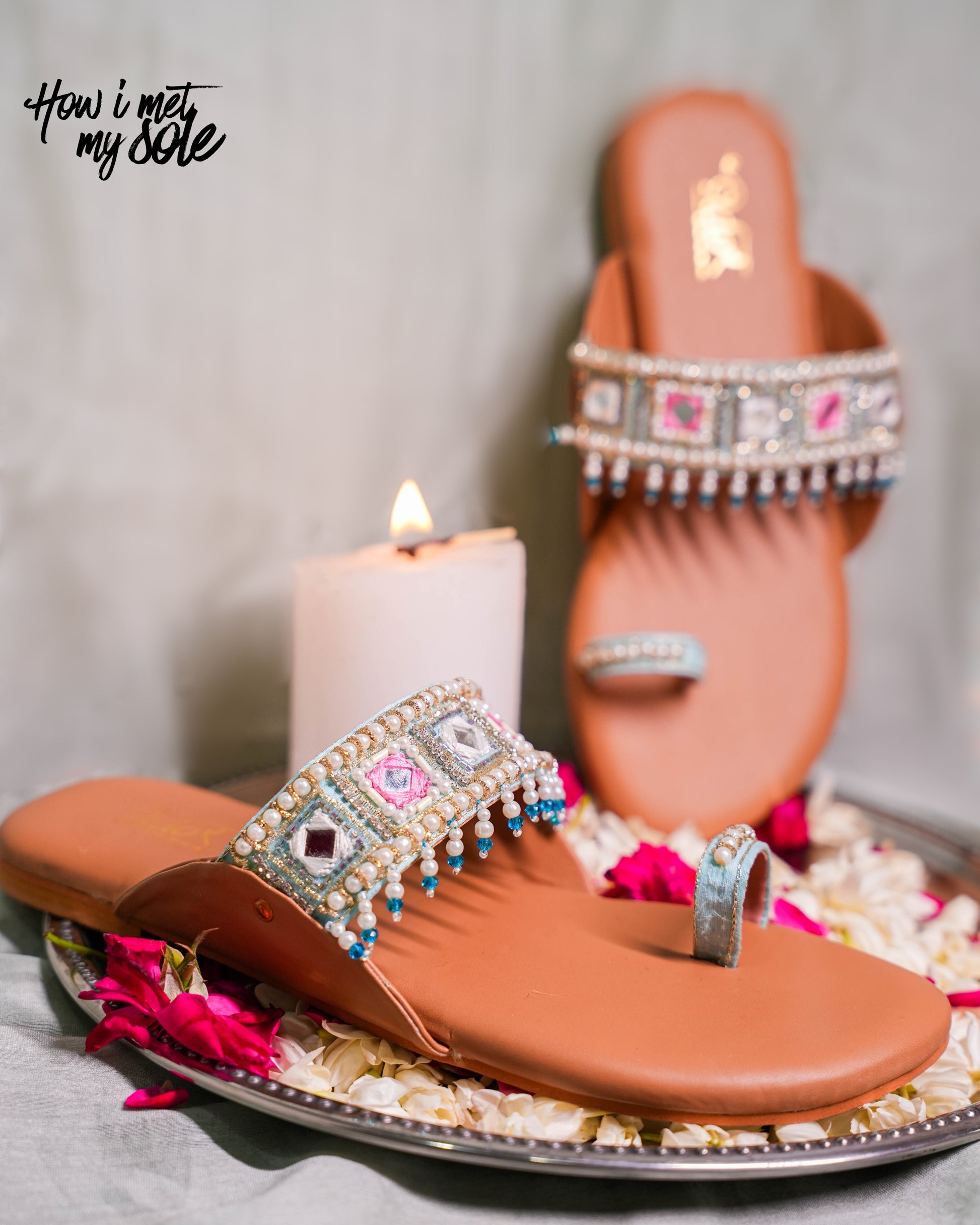 Pair of ornate brown sandals with decorative straps on a gray surface with a candle and flowers.